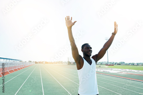 An African American man in a white T shirt happily runs under the sun lifting the top after winning the race