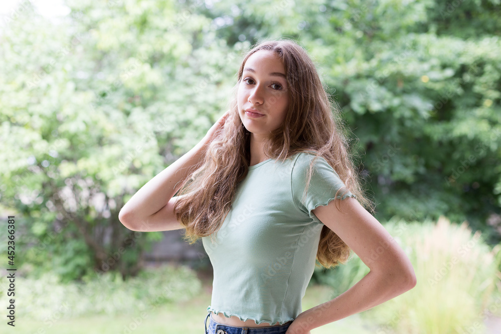 Medium horizontal view of beautiful teenaged girl with long dirty blond curly hair dressed in green shirt seen standing in front of a garden in summer Medium horizontal view of beautiful teenaged girl with long dirty blond curly hair dressed in green shirt seen standing in front of a garden in summer