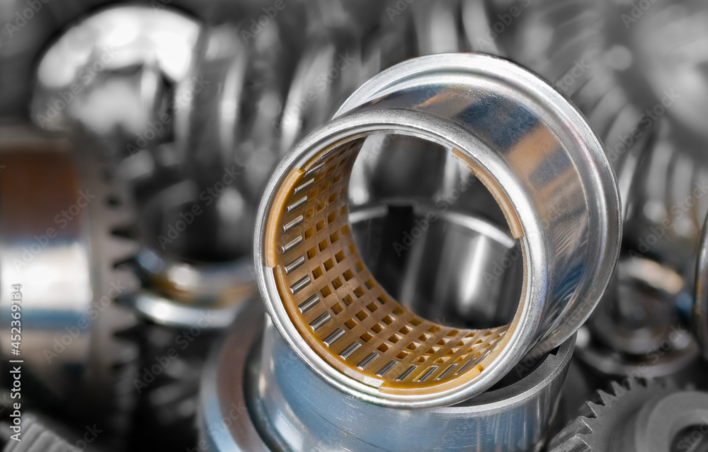 Closeup of needle roller bearing with plastic cylindric cage in steel bushing. Rolling metal part on blur background of pile of hardware components as gear wheel cogs or coil springs. Selective focus. Closeup of needle roller bearing with plastic cylindric cage in steel bushing. Rolling metal part on blur background of pile of hardware components as gear wheel cogs or coil springs. Selective focus.