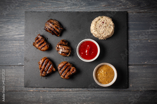 Grilled pork medallions with ketchup and mustard on dark wooden table.