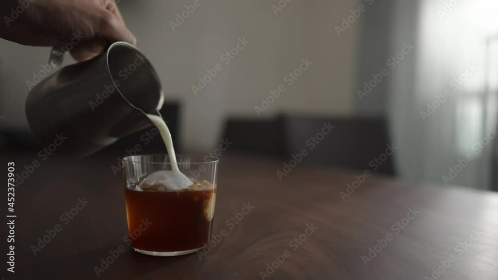 Slow motion pour milk into cold brew coffee over ice ball in tumbler glass on walnut table Slow motion pour milk into cold brew coffee over ice ball in tumbler glass on walnut table