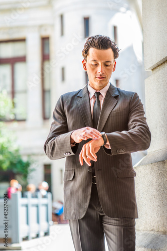 Dressing formally,  holding a watch in his wrist, a young businessman is standing outside an office building, looking down and checking time..