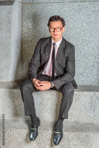 Dressing in a black striped jacket, tie, pants, leather shoes, wearing glasses and a watch, arms resting on laps,  a young professor is sitting outside, relaxing and thinking. © Alexander Image