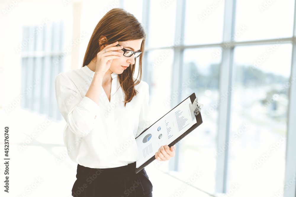 Beautiful female specialist with clipboard standing in modern office and smiling charmingly. Working on design, data analysis, plan strategy. Business people concept Beautiful female specialist with clipboard standing in modern office and smiling charmingly. Working on design, data analysis, plan strategy. Business people concept