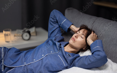 Depressed mature woman in pajamas lying in bed and touching head with hands. Caucasian brunette unable to sleep at night because of insomnia.