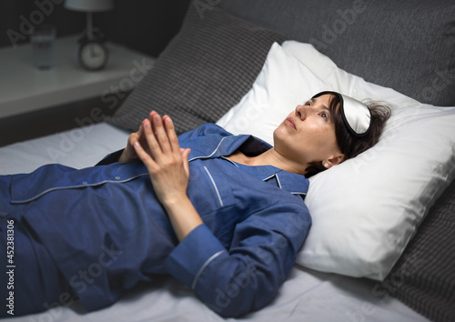 Pensive mature woman in sleep mask and pajamas lying on white soft linen and keeping eyes open during night. Concept of insomnia, problems and sleep disorder.