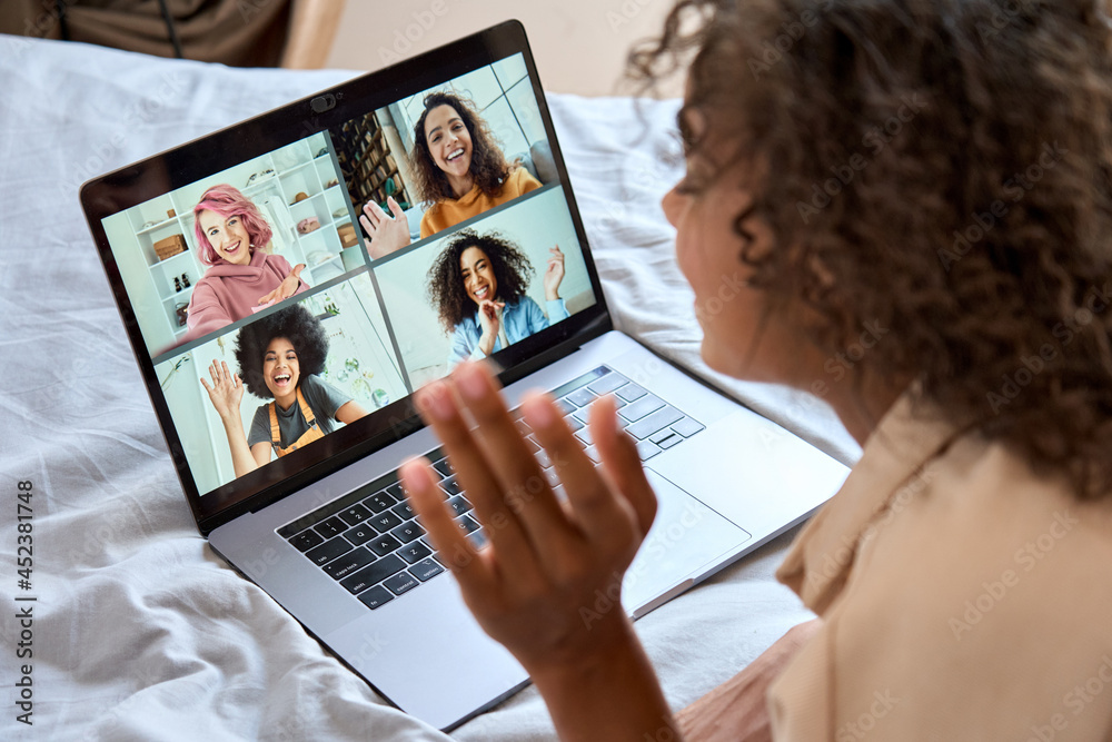 © insta_photos - Young African American teen girl lying on bed having virtual meeting waving hand chatting with friends during online video call at home having fun using laptop computer. Over shoulder view
