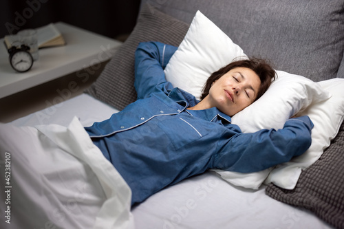 Pretty mature woman in blue pajamas resting on comfy bed with white soft pillows and blanket. Caucasian female with brown hair sleeping peacefully at night.
