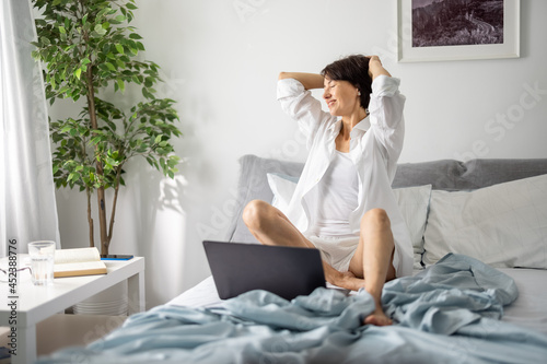 Pleasant mature woman resting on bed during morning time and using wireless laptop. Positive caucasian lady in sleepwear enjoying carefree moments at home.