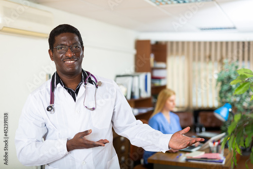 Smiling african american doctor wearing white coat with phonendoscope on his neck welcoming to medical office, making inviting gesture with hand