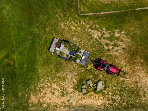 A red tractor pulls a trailor with plants on it through a patchy green grass field on a sunny clear summer day.  Aerial perspective looking down as the photograph was shot from an drone.