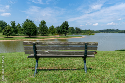 An old deteriorating wood and metal park bench in a grass field on a sunny summer day surrounded by tall trees.