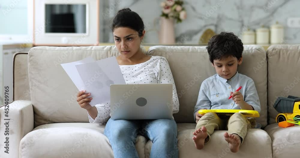 Busy concentrated Indian entrepreneur woman do remote paperwork accounting job use laptop working sit on sofa with little son drawing on magnetic erasable board. Quarantine at home with kids concept