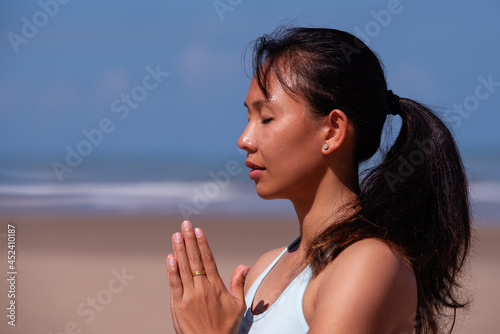 people, fitness, sport and healthy lifestyle concept - close up portrait of young asian woman making meditation on tropical beach with blue sky background
