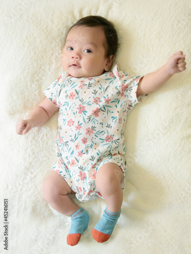 Top view of cute little Asian baby girl, wearing a pretty floral dress, lying on white bed and looking at the camera