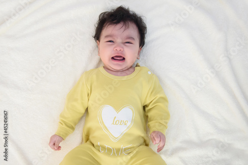 Top view of a cute little Asian baby girl wearing yellow dress, lying on the bed, sad and crying