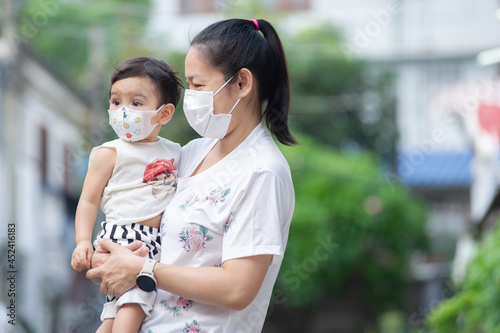Asian mother wearing white face mask holding her little toddler baby girl who is her daughter wear baby face mask, they looking out, COVID-19 concept