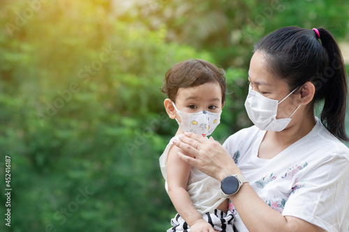 Asian mother wearing white face mask holding her little toddler baby girl who is her daughter wear baby face mask, mom look at baby, COVID-19 concept