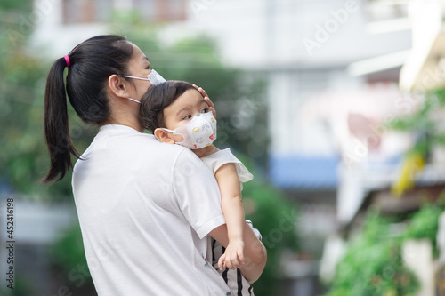 Asian mother wearing white face mask holding her little toddler baby girl who is her daughter wear baby face mask, looking out, COVID-19 concept