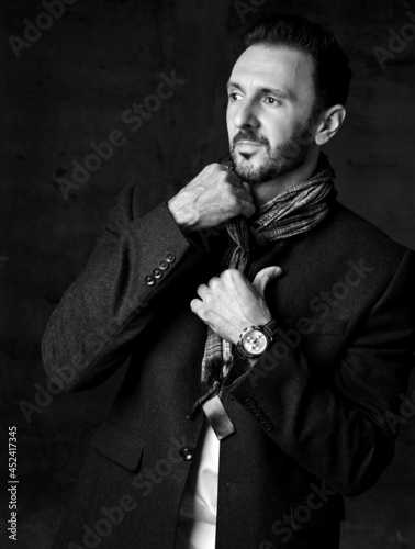 Black and white. Elegant unshaved adult man in black jacket stands putting on, taking off his stylish scarf and looks aside over dark background