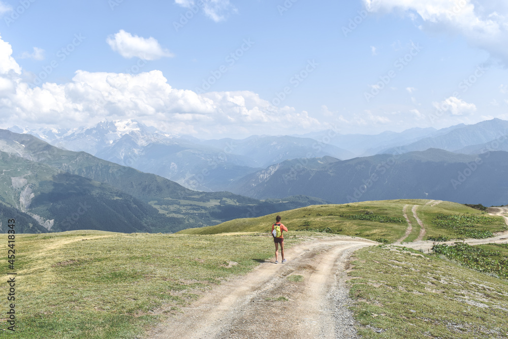 Female hiker on a trail in Svaneti region, Georgia, Asia. Summer mountain landscape with snowcapped mountains in the background. Blue sky with clouds above. Georgian tourist destination