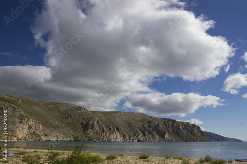 a landscape of blue-gray water with a sandy beach against the background of brown-green rocky mountains and a blue-blue sky with a large white cloud