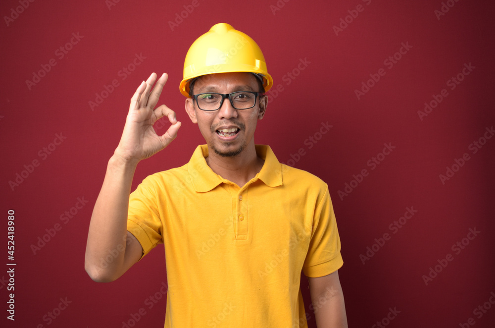 Handsome Asian worker man wearing helmet and glasses smiling and shows okay gesture against red background.