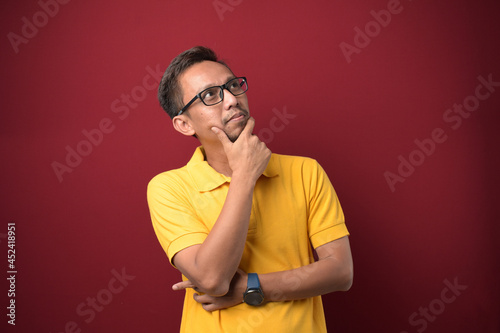Asian man wearing glasses thinking and looking up over on yellow background with hand on chin.