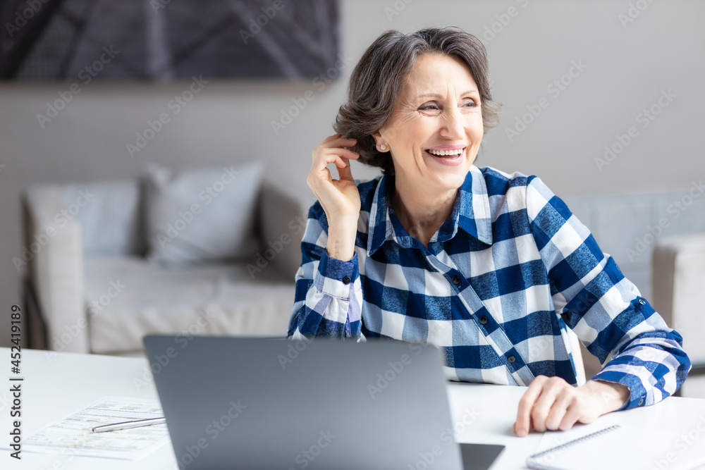 Portrait of a happy business aged woman with laptop computer sitting at a desk in an office or at home looks away from the window and smiles Portrait of a happy business aged woman with laptop computer sitting at a desk in an office or at home looks away from the window and smiles
