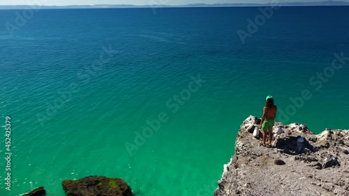 Mexican Man Standing On Top Of Coastal Cliff Overlooking Wide Blue Ocean In Summer. Playa Balandra In Baja California Sur, Mexico. aerial drone orbit photo