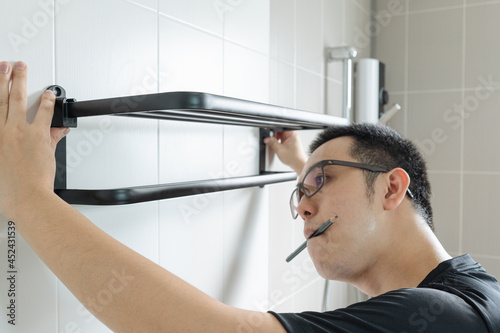 Asian man preparing to install a towel rail on the bathroom wall He put the rack against the wall and used a pencil to mark where to drill.