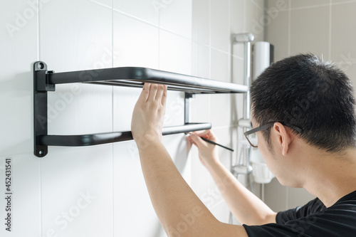 Asian man preparing to install a towel rail on the bathroom wall He put the rack against the wall and used a pencil to mark where to drill.