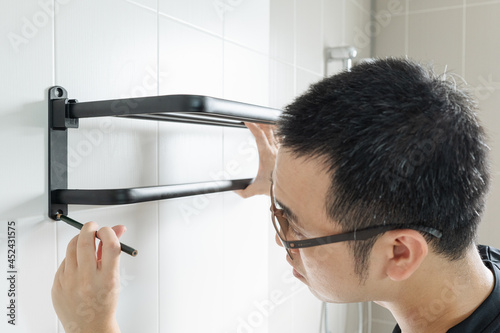 Asian man preparing to install a towel rail on the bathroom wall He put the rack against the wall and used a pencil to mark where to drill.