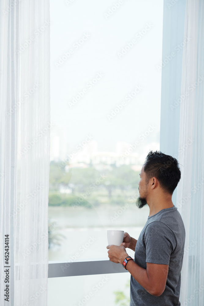 Pensive man drinking morning coffee and looking outdoors through big apartment window Pensive man drinking morning coffee and looking outdoors through big apartment window