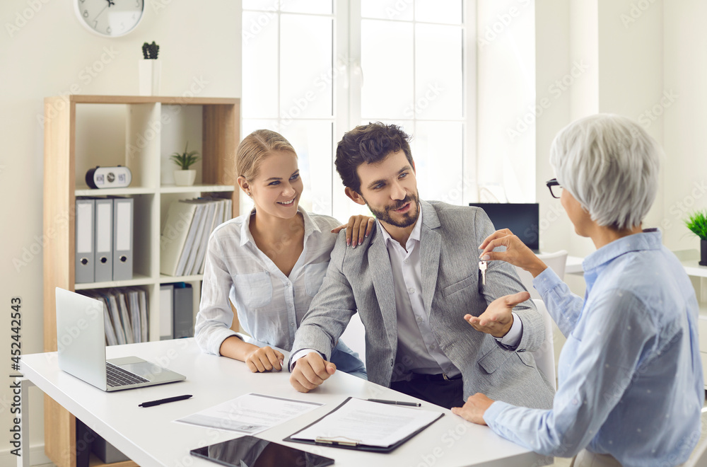 Obraz premium Senior realtor gives key to new house or apartment to happy young married couple after they have signed agreement. Concept of real estate, mortgage, rent, lease, or buying your own property
