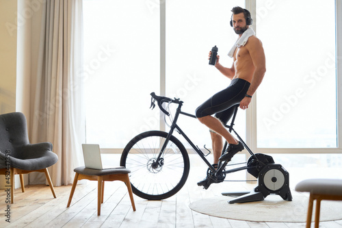 Athletic young man in headphones riding stationery bike at home