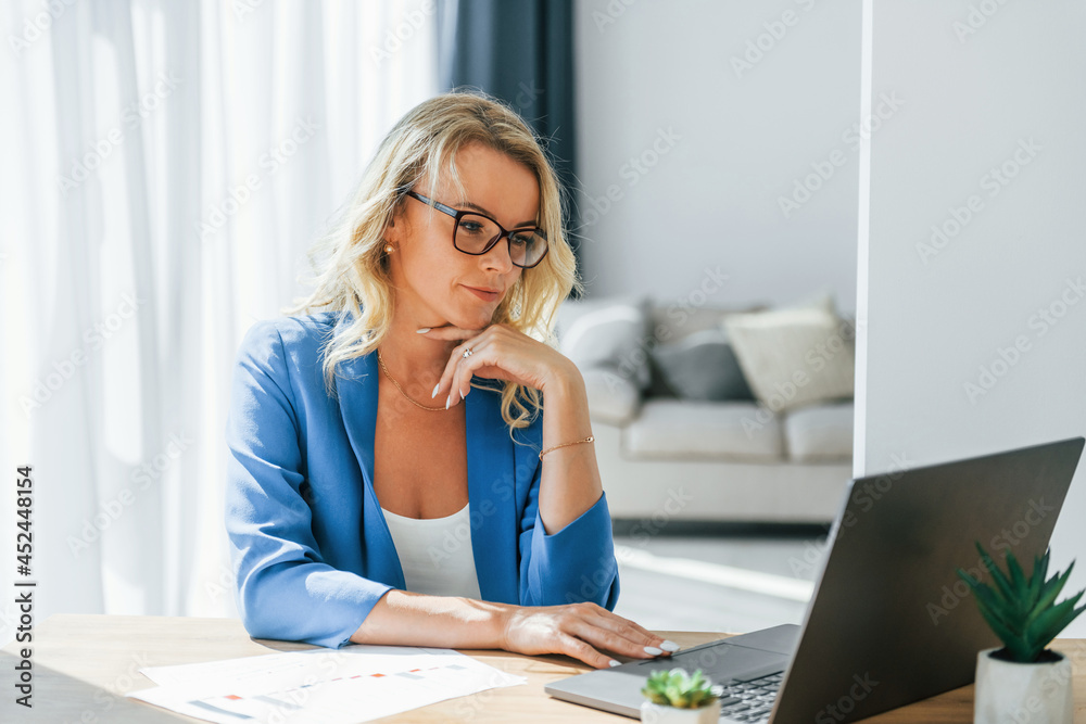 Sits by table with laptop. Woman in casual clothes is indoors at home Sits by table with laptop. Woman in casual clothes is indoors at home