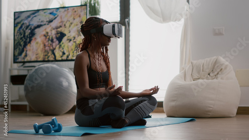Black trainer woman wearing virtual reality headset while sitting on yoga map in lotus position during meditation workout class in living room. Athetic adult enjoying healthy lifestyle training body photo
