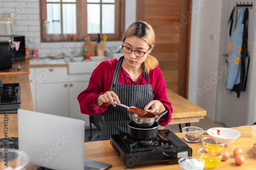 Beautiful asian woman learn to cook chocolate cake, video recipes on a laptop in the kitchen, online learning cooking at home concept