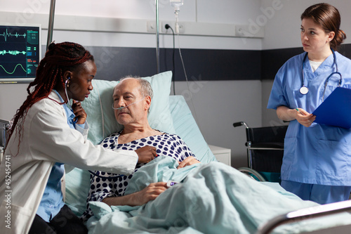 Senior man breathing with help from oxygen tube laying in hospital bed, listening african american doctor explaying diagnosis treatmant holding pills bottle for recovery. photo