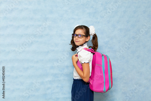 Billede på lærred Funny dark-haired girl in glasses in school clothes with a backpack on her back on a blue background