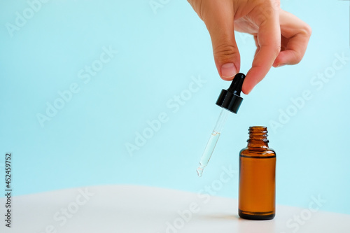 Female hand holds the essential oil falling from the amber glass dropper. Bottle of cosmetic oil with a pipette. Copy space. Light blue background.