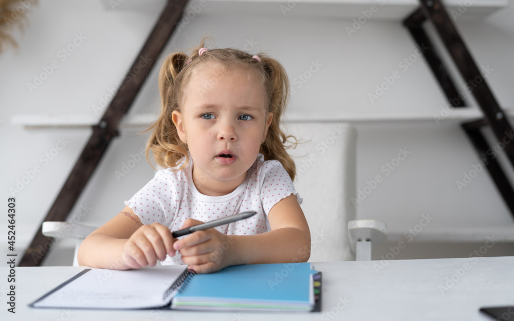 pretty little elementary school girl doing her homework at home while sitting at her desk with a notebook and pen for drawing and handwriting. Pupil school is studying remotely.