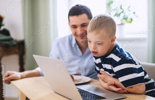 Father with happy down syndrome son indoors at home, using laptop.
