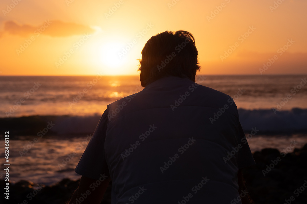 Rear view of mature man at sea looking at the horizon and at the orange sunset light Rear view of mature man at sea looking at the horizon and at the orange sunset light