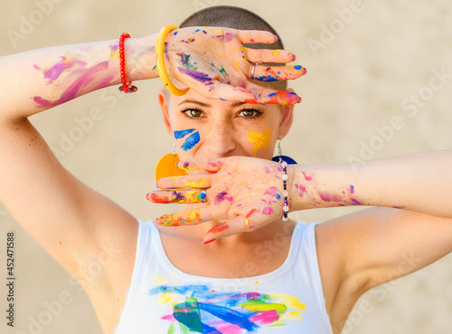 Playful portrait of a young gorgeous female painter artist, with hands covered in paint, looking and smiling at camera through her fingers. Creativity and individuality concept.