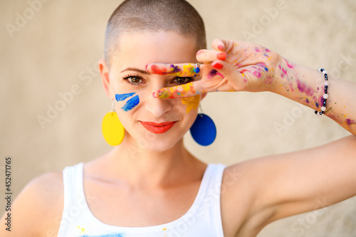 Playful portrait of a young gorgeous female painter artist, with hands covered in paint, looking and smiling at camera through her fingers. Creativity and individuality concept. photo