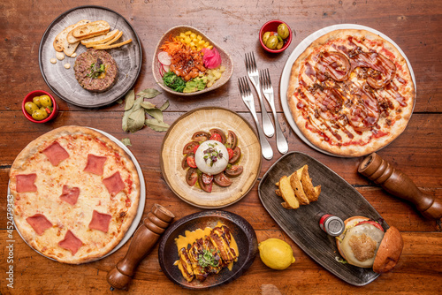 Top view image of pizzas and Mediterranean food dishes. Beef burger, burrata salad, guanciale, noturego salmon, poke bowl, Spanish olives and pepper shakers with lemon.