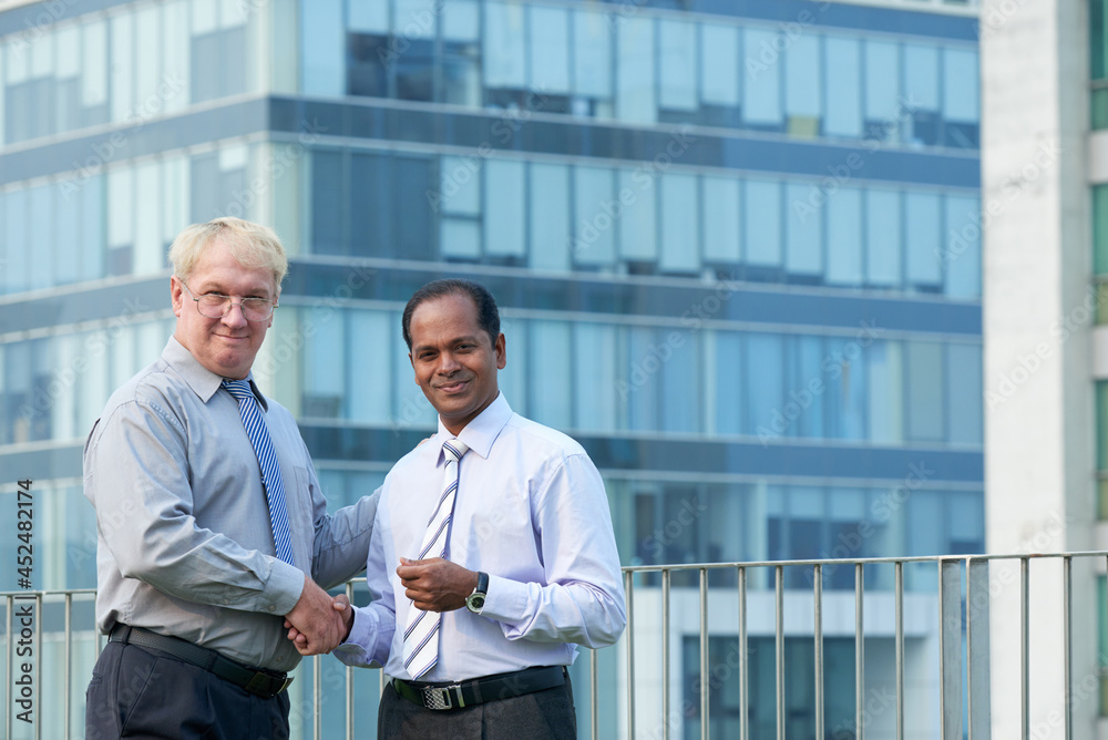 Positive mature entrepreneur shaking hand of investor after meeting, office building or skyscraper in background Positive mature entrepreneur shaking hand of investor after meeting, office building or skyscraper in background
