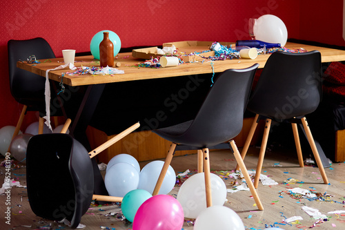 After party chaos, messy in livving room at home, table with pizza and champagne glasses covered with confetti and ballons, chair on the floor at morning after party celebration. photo